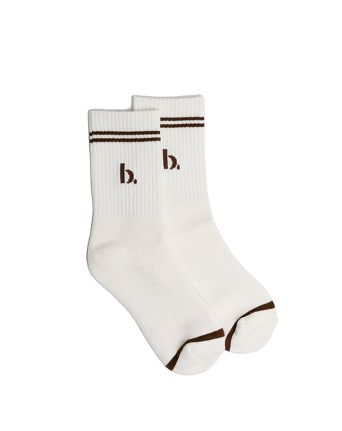 A pair of white ribbed crew socks with brown stripes on the cuff, a brown embroidered lowercase 'b.' logo on the front, and brown trim on the toes, shown against a plain white background.